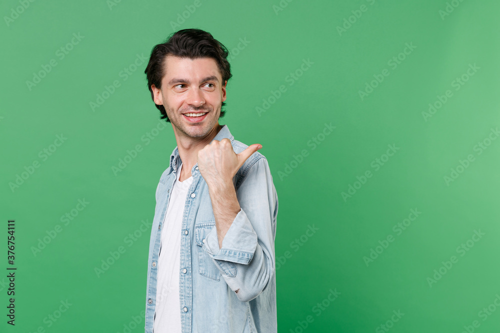 Side view of smiling young brunet man 20s wearing casual clothes white t-shirt denim shirt posing stand pointing thumb aside on mock up copy space isolated on green color background studio portrait.