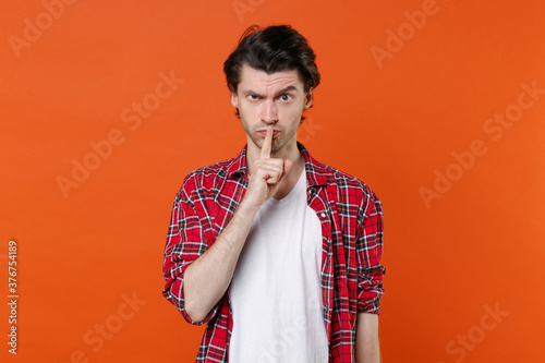 Secret young brunet man 20s wearing white t-shirt red checkered shirt posing saying hush be quiet with finger on lips shhh gesture looking camera isolated on orange color background studio portrait.