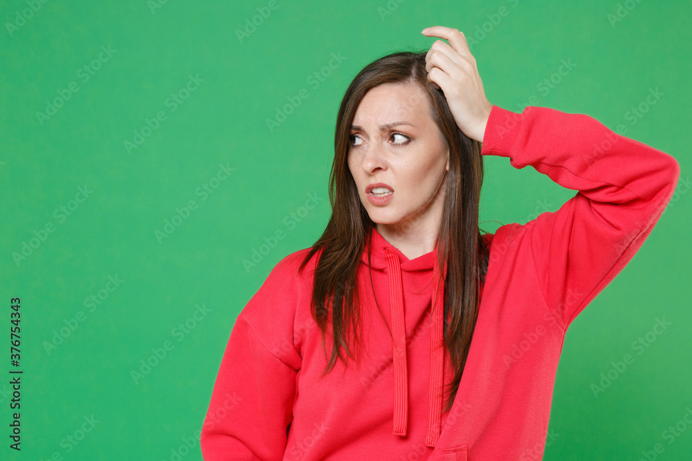 Preoccupied puzzled concerned young brunette woman 20s wearing bright red casual streetwear hoodie posing put hand on head looking aside isolated on green color wall background studio portrait.