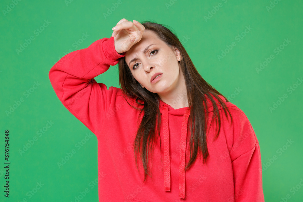 Exhausted tired displeased sick young brunette woman 20s wearing bright red casual streetwear hoodie posing put hand on head looking camera isolated on green color wall background studio portrait.