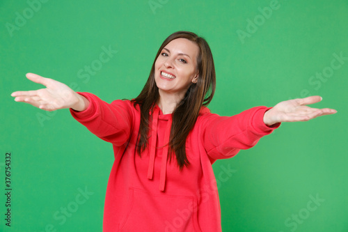Smiling joyful beautiful young brunette woman 20s wearing bright red casual streetwear hoodie posing reach out stretch hands looking camera isolated on green color wall background studio portrait.