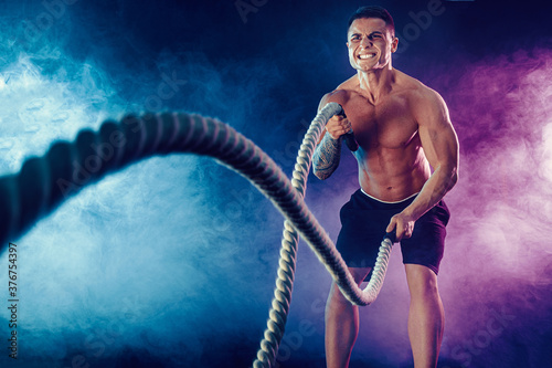 Athletic looking bodybulder work out with battle rope on dark studio background with smoke. Strength and motivation.