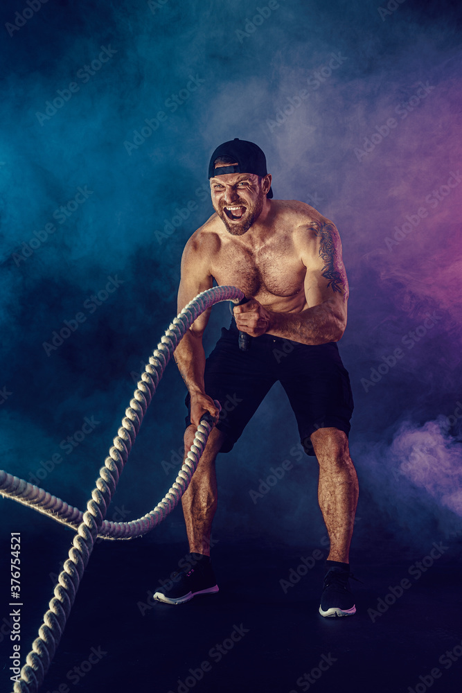 Bearded athletic looking bodybulder work out with battle rope on dark studio background with smoke. Strength and motivation.