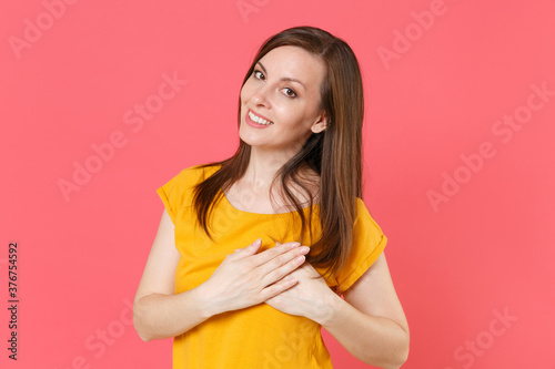 Smiling pleasant beautiful young brunette woman 20s wearing yellow casual t-shirt posing standing holding hands folded on heart chest looking camera isolated on pink color background studio portrait.