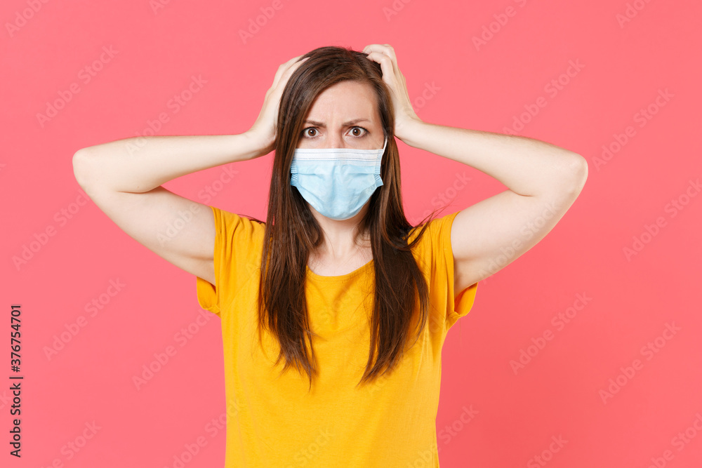 Worried young woman in yellow t-shirt sterile face mask to safe from coronavirus virus covid-19 2019-ncov during pandemic quarantine put hands on head isolated on pink wall background studio portrait.