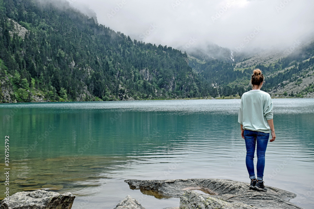 Fototapeta premium A woman stands in a rock in a green mountain landscape with a large lake of transparent water, trees and clouds