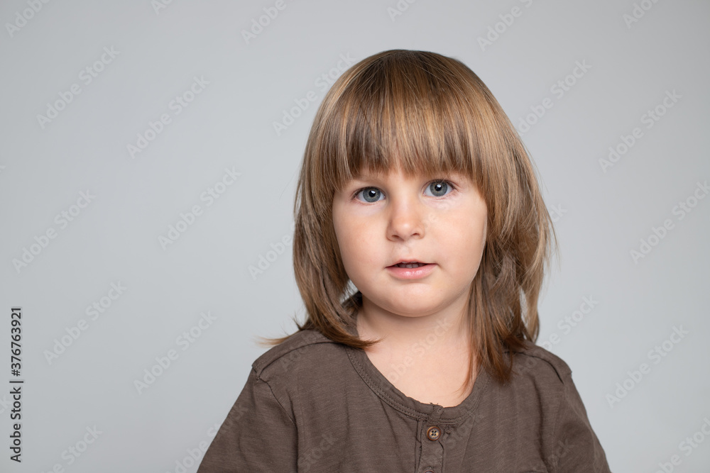 Portrait of cute boy with long thick hair. The child has blue eyes, brown hair, thick bangs. He is in a brown t-shirt. Cheerful kid smiling. Caucasian. Looks at the camera. Studio, gray background. Portrait of cute boy with long thick hair. The child has blue eyes, brown hair, thick bangs. He is in a brown t-shirt. Cheerful kid smiling. Caucasian. Looks at the camera. Studio, gray background.
