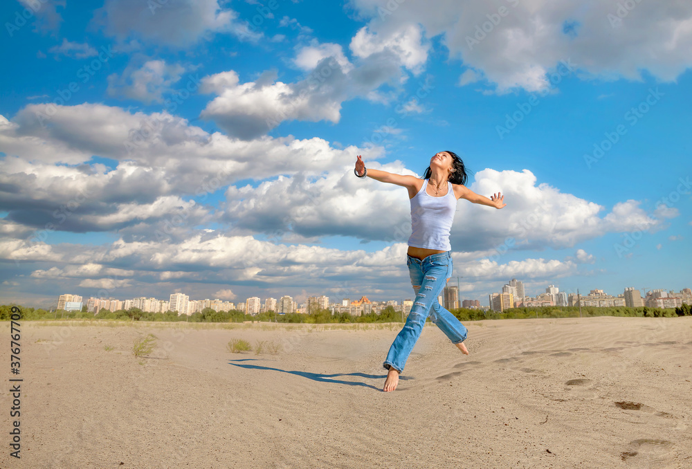 Fototapeta premium Joyful young woman in blue jeans jumping on sandy beach outside city