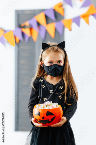 A girl in a Halloween costume with a mask on is holding a pumpkin filled with treats during the covid19 pandemic at a fall festival.