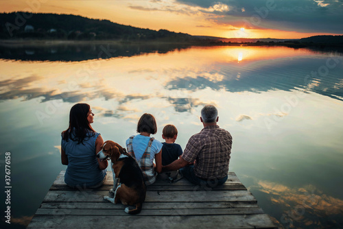 family of four people sitting on the shore on a wooden bridge of a large lake in summer and watching the beautiful sunset with dog back view