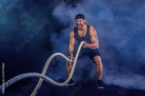 Bearded athletic looking bodybulder work out with battle rope on dark studio background with smoke. Strength and motivation.