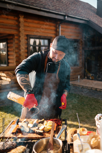 A tattooed chef pouring oil on vegetables and meat while cooking steak in nature. Concept of the correct cooking of meat.
