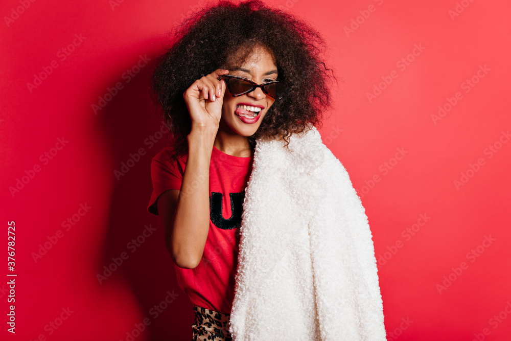 Glad african woman in sunglasses making faces on red background. Indoor portrait of inspired brunette girl in white autumn coat. Glad african woman in sunglasses making faces on red background. Indoor portrait of inspired brunette girl in white autumn coat.