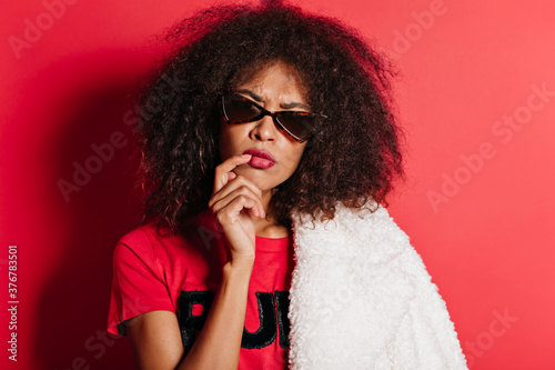 Funny pretty woman with curly hairstyle thinking about something. Indoor portrait of lovable african lady isolated on bright red background.