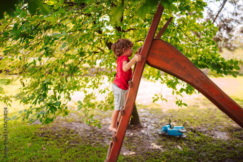 A beautiful little caucasian boy in a red T-shirt is playing on a wooden slide in the park.