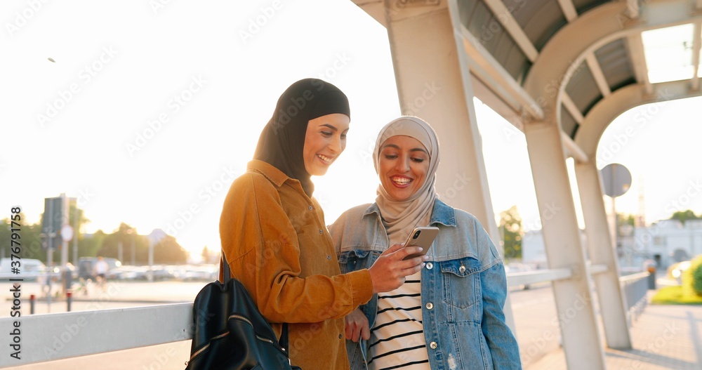 Two Arabian pretty females in headscarves standing outdoors watching something on mobile phone. Muslim beautiful women using smartphone and talking. Tapping and browsing.