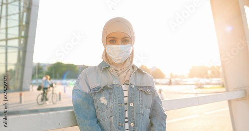 Portrait shot of beautiful happy young Arabian woman in traditional headscarf and medical mask looking at camera in sunlight. Sunny summer outdoor. Muslim joyful female in hijab. Islamic girl in city. photo