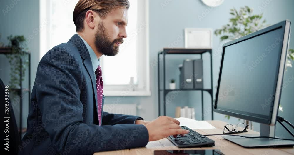 Side view of bearded company worker in business suit sitting at table and working on computer. Confident man using modern technology for work at office. Side view of bearded company worker in business suit sitting at table and working on computer. Confident man using modern technology for work at office.