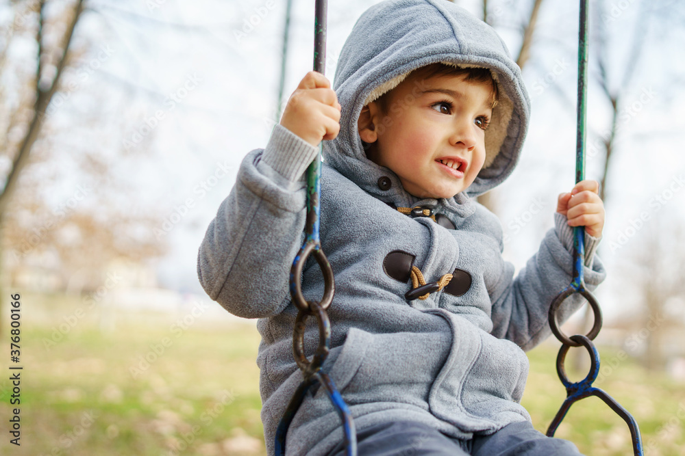 Side view on small caucasian boy in winter coat swinging in park having fun alone in autumn day - copy space leisure activity growing up concept Side view on small caucasian boy in winter coat swinging in park having fun alone in autumn day - copy space leisure activity growing up concept