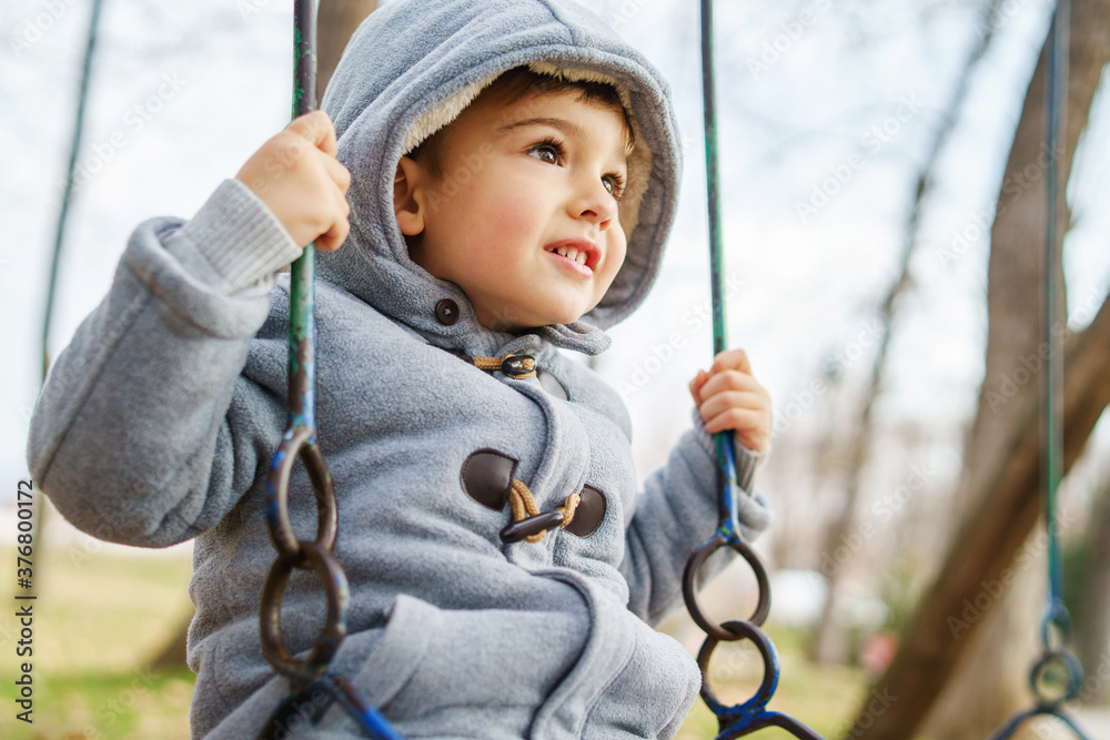 Side view low angle on small caucasian boy in winter coat swinging in park having fun alone in autumn day - leisure activity growing up concept Side view low angle on small caucasian boy in winter coat swinging in park having fun alone in autumn day - leisure activity growing up concept