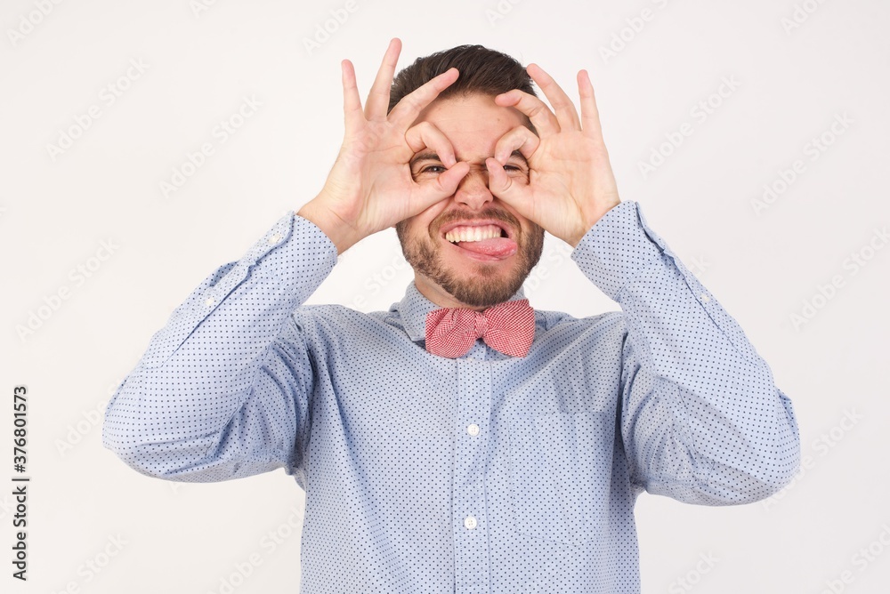 European man dressed in formal shirt and bow tie poses against white background doing ok gesture like binoculars sticking tongue out, eyes looking through fingers. Crazy expression. European man dressed in formal shirt and bow tie poses against white background doing ok gesture like binoculars sticking tongue out, eyes looking through fingers. Crazy expression.