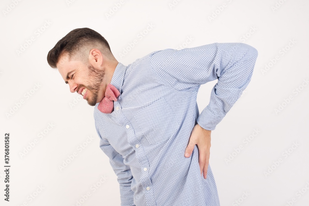 European man dressed in formal shirt and bow tie poses against white background Suffering of backache, touching back with hand, muscular pain European man dressed in formal shirt and bow tie poses against white background Suffering of backache, touching back with hand, muscular pain