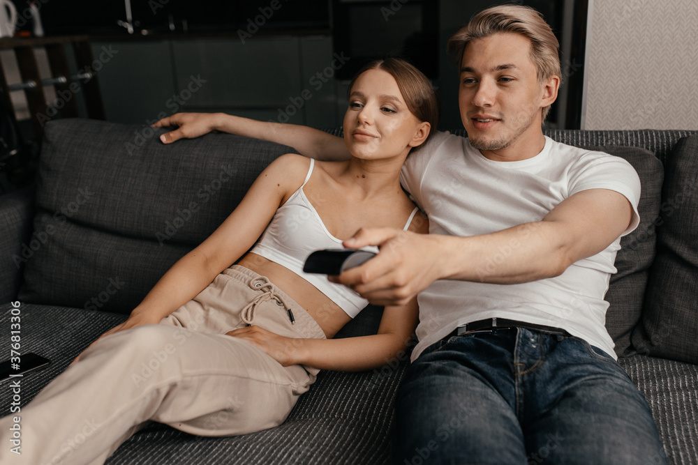 A man and a beautiful woman are sitting on the couch in the apartment and watching TV. Family everyday life. A man and a beautiful woman are sitting on the couch in the apartment and watching TV. Family everyday life.