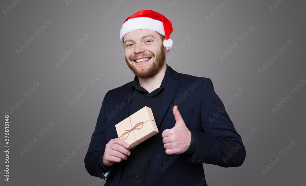 Bearded entrepreneur wearing a santa hat is posing in a black suit with a present gesturing the like sign on a gray studio wall Bearded entrepreneur wearing a santa hat is posing in a black suit with a present gesturing the like sign on a gray studio wall