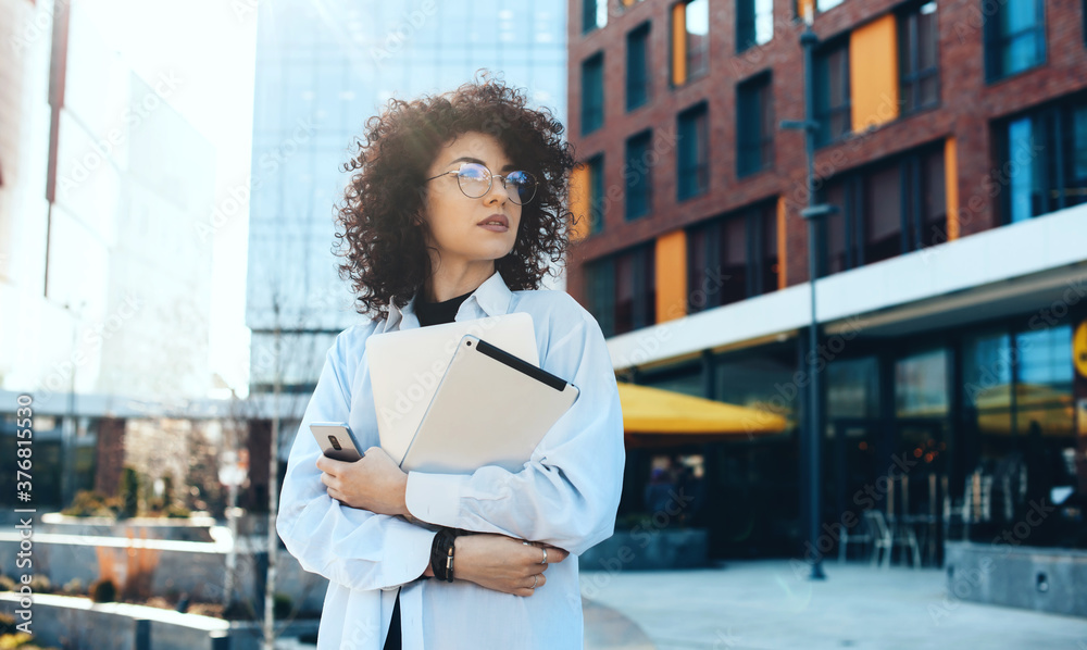 Front view photo of a modern it woman with curly hair and eyeglasses posing in the city with a tablet and laptop while waiting for somebody