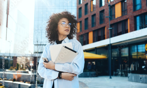 Front view photo of a modern it woman with curly hair and eyeglasses posing in the city with a tablet and laptop while waiting for somebody