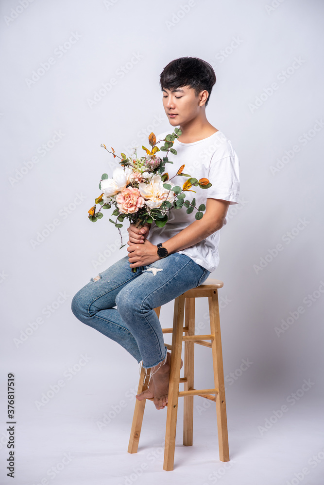 A young man in a white t-shirt sits on a high chair and holds flowers. A young man in a white t-shirt sits on a high chair and holds flowers.