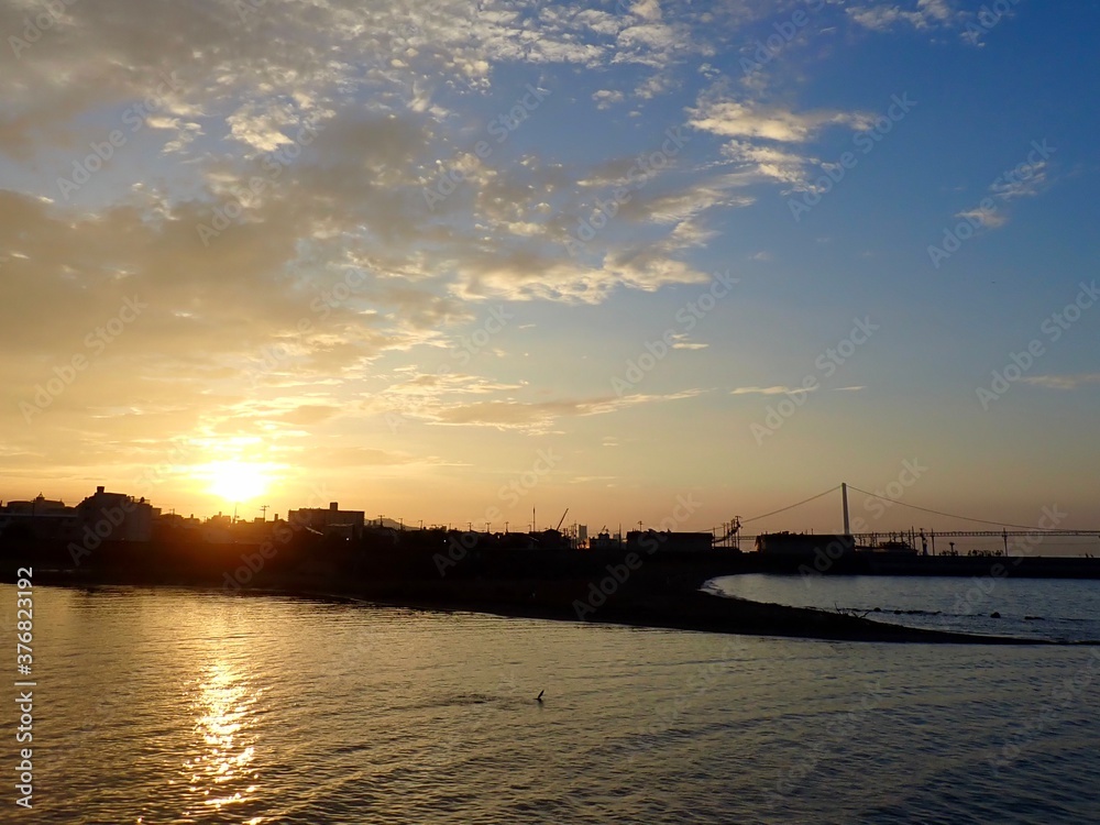 白い雲が浮かぶ青空に上ってくる朝日とその光が映りオレンジに輝く河口の水面