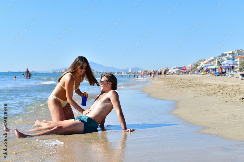 Young couple smiling while the woman rubs the man with sunscreen at the seashore. New travel and healthcare concept. Young couple smiling while the woman rubs the man with sunscreen at the seashore. New travel and healthcare concept.