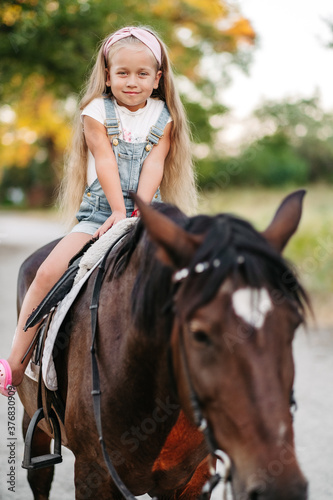 Little blonde girl with long hair rides a horse in the park at sunset in autumn. Autumn horse ride. Friendship of a girl and a horse.