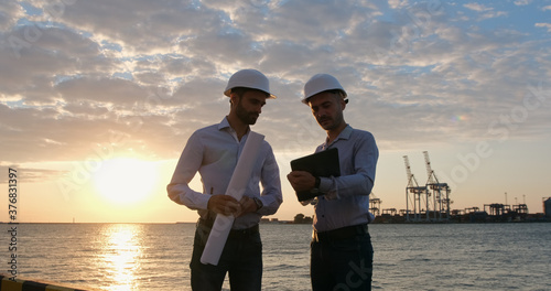 Two engineers in white protective helmets work with a plan on a tablet in front of the seaport in the early morning.