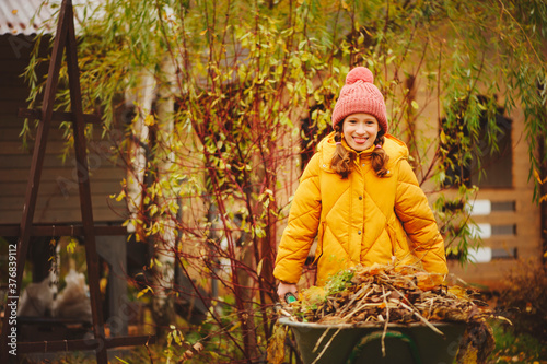 Happy child girl working in autumn garden, helps to remove fall leaves from lawn