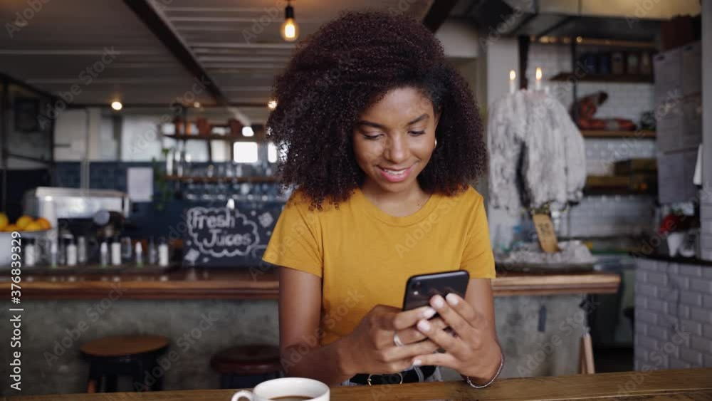 African American smiling female typing to boyfriend on smartphone sitting in trendy cafe next to hot cup of coffee. African American smiling female typing to boyfriend on smartphone sitting in trendy cafe next to hot cup of coffee.