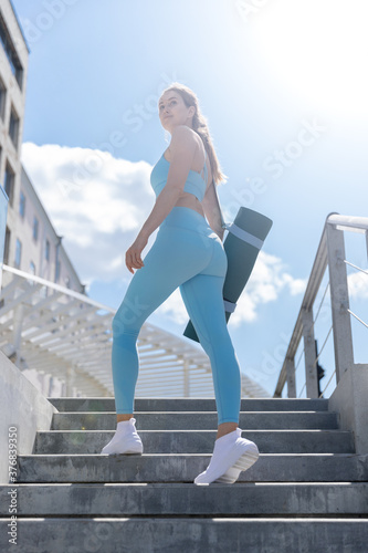Young smiling female in stylish sportswear holding yoga mat walks up stairs for workout in city. 