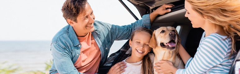 Horizontal crop of family with golden retriever standing near auto on beach