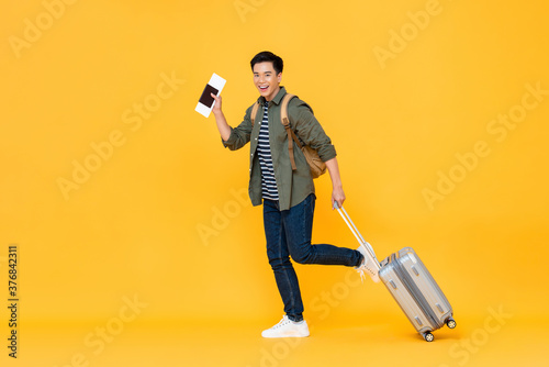 Full length portrait of smiling happy handsome young Asian man tourist with passport and luggage ready to travel on vacations isolated in yellow studio background photo