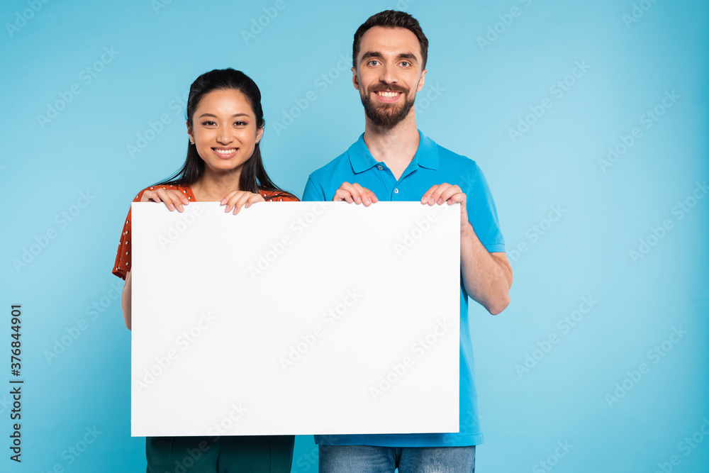 young interracial couple looking at camera while holding empty board on blue young interracial couple looking at camera while holding empty board on blue