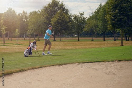 Man playing golf going to the green with his putter that he has just taken from his golf bag with a sand hazard or bunker in the foreground