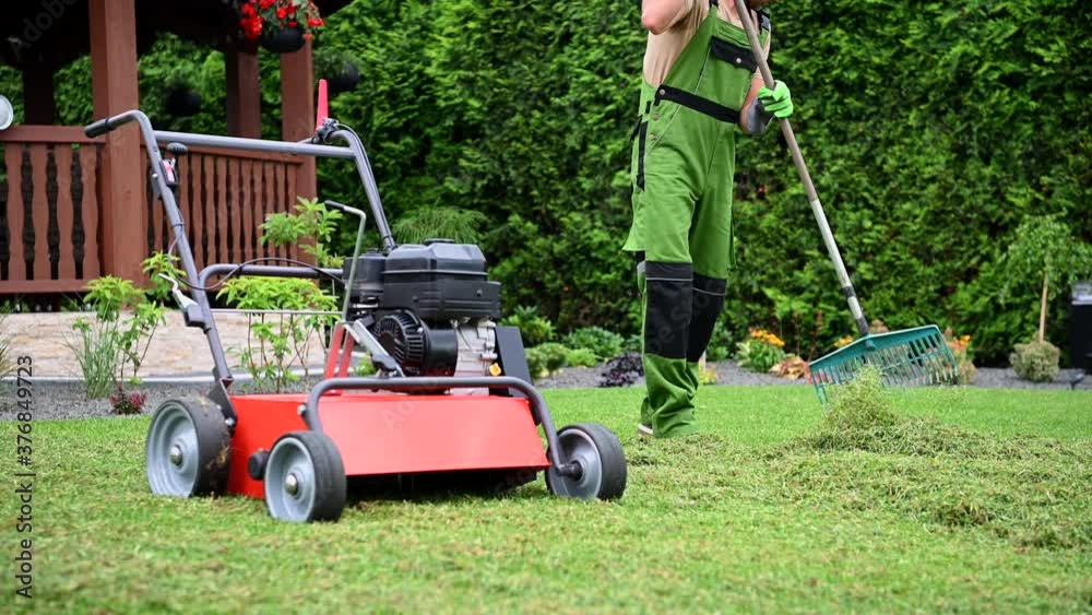 Male Worker Tidying Up Grass Clippings After Mowing Residential Yard Area. Garden Maintenance With Mower And Rake. Male Worker Tidying Up Grass Clippings After Mowing Residential Yard Area. Garden Maintenance With Mower And Rake.