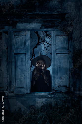 Scary halloween witch standing in old damaged wood window with wall over cross, church, birds, dead tree and spooky cloudy sky, Halloween mystery concept