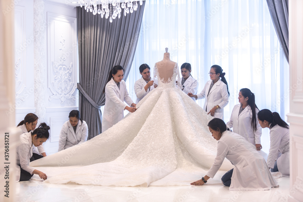 Team of female dressmaking workers preparing a large haute couture wedding dress on a mannequin in a bridal design studio. - obrazy, fototapety, plakaty 