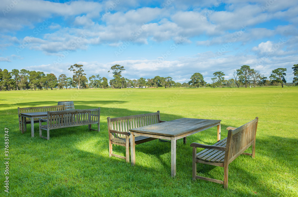 Two wooden dining tables and some sitting benches on a vast grass field with large open space. Concept of relaxation, refreshment and outdoor rest area with a view. Background texture with copy space.