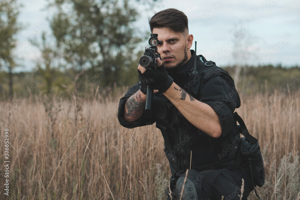 Fototapeta premium Young soldier in black uniform sitting and aiming with an assault rifle.