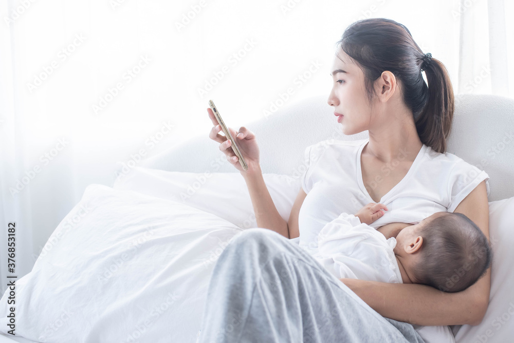 Mother breastfeeding her newborn baby beside window. Milk from mom’s breast is a natural medicine for children. Young woman feeding baby. Mom using the phone. Mother breastfeeding her newborn baby beside window. Milk from mom’s breast is a natural medicine for children. Young woman feeding baby. Mom using the phone.