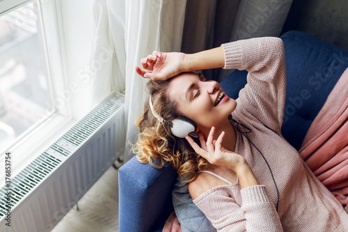 Indoor winter photo of dreamy woman in cozy knitted loungewear enjoying lovely music by earphones.