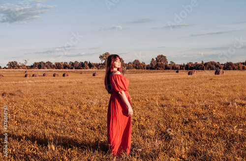 female model plus size in red dress on a field with haystacks, a beautiful young woman with brown hair, harvest concept © klavdiyav